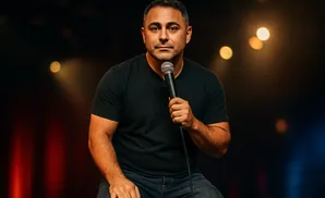 A man in a black t-shirt and jeans holds a microphone on a stage with blurred lights.