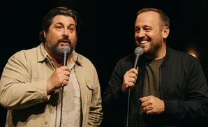 Two men on stage, holding microphones and laughing, one points at the camera.