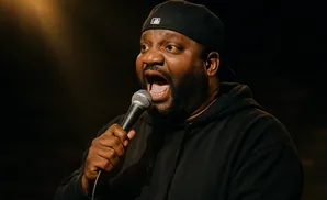 Man in a black hoodie and baseball cap speaks into a microphone.