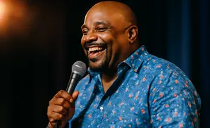 A bald Black man in a blue floral shirt laughs while holding a microphone.