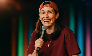 A smiling woman in a baseball cap holds a microphone on a stage with colorful lights.