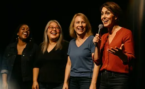 Four women on a stage, three laughing and one speaking into a microphone.