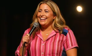 A woman with blonde hair smiles into a microphone while wearing a pink and red striped shirt.