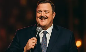 Billy Gardell smiles as he holds a microphone on stage.