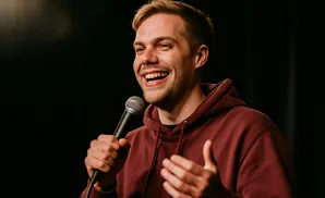 A young man with blonde hair and a beard laughs while holding a microphone and gesturing with his hand.