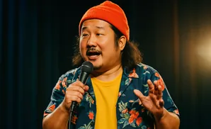Comedian Bobby Lee wears an orange beanie and floral shirt while holding a microphone and speaking on stage.