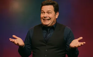 A man in a vest and tie gestures with his hands against a colorful background.