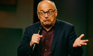 Man in a suit and glasses makes a surprised face while holding a microphone.