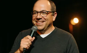 Man in glasses and a gray sweater smiles while holding a microphone.