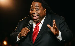 A Black man with dreadlocks wearing a suit and red tie holds a microphone and gestures with his hands.