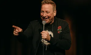 A man in a black bomber jacket with a rose emblem smiles and points while holding a microphone.