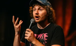 A man with curly hair and a baseball cap tells a joke into a microphone.