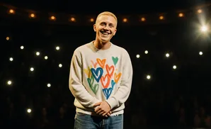 A smiling man with a buzz cut wears a cream sweater with colorful hearts and blue jeans on stage.