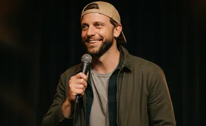 Comedian Andrew Schulz in a tan baseball cap and olive jacket, holding a microphone and smiling.