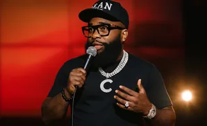 Comedian holding microphone, wearing glasses, black cap, and bling.