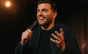 A smiling man holds a microphone and gestures with his hand while on stage.