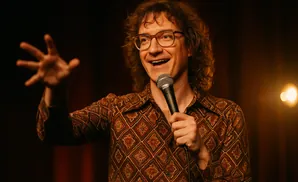 A smiling man with curly hair and glasses, wearing a patterned shirt, holds a microphone and gestures with his hand.