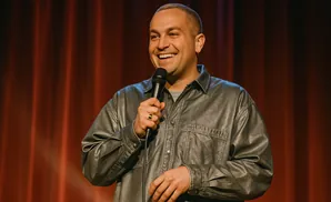A man in a gray shirt and jacket smiles while holding a microphone on a red stage.