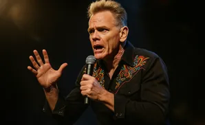 Comedian Brian Posehn performs onstage with a microphone in a black shirt with colorful embroidery.