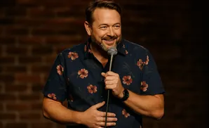 A man wearing a dark blue floral shirt smiles and holds a microphone while standing in front of a brick wall.