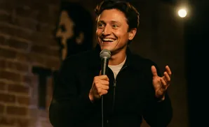 A man in a black jacket smiles widely while holding a microphone, with a spotlight and brick wall in the background.