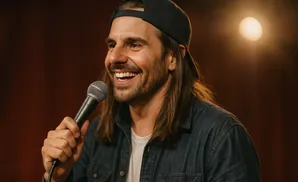 A man with long hair and a baseball cap smiles while holding a microphone.