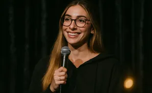 A smiling woman wearing glasses and holding a microphone stands in front of a dark background.