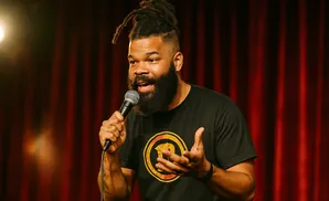 Man with dreadlocks and a beard speaks into a microphone on a stage with red curtains.