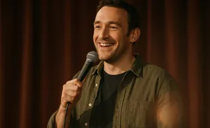 A man in a green shirt and black t-shirt holds a microphone and smiles in front of a brown curtain.