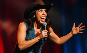 A woman in a cowboy hat and denim vest holds a microphone and gestures with her hand.