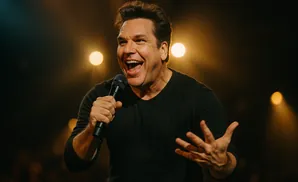 Comedian laughing into a microphone with his hand gesturing, on a dimly lit stage.
