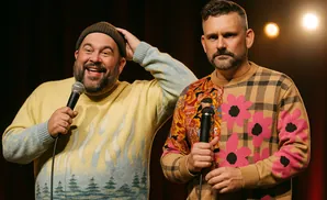Two men with microphones stand on stage, one wearing a beanie and a sweater with a flame and tree design, the other a sweater with plaid and flower patt…