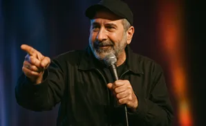 A man in a black jacket and baseball cap holds a microphone and points.