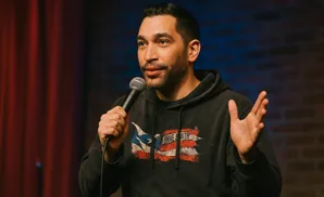 A man with a beard and a dark hoodie with a distressed American flag design holds a microphone and gestures with his hand.