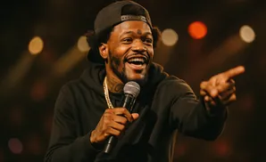 A man in a black hoodie and baseball cap laughs while holding a microphone and pointing.