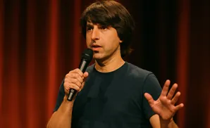 Comedian David Spade holding a microphone and gesturing with his left hand.