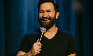 Man in a black v-neck shirt smiles and holds a microphone, with a dark blue curtain behind him.