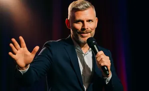 A man in a suit holds a microphone and waves his hand.