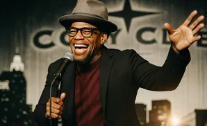 A Black man in a fedora and suit laughs while holding a microphone on stage, with a city skyline backdrop.