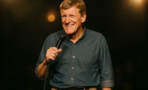 A smiling man in a denim shirt holds a microphone while speaking on a stage.