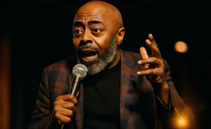 A bald Black man with a gray beard speaks into a microphone, wearing a plaid jacket and gesturing with his hand.
