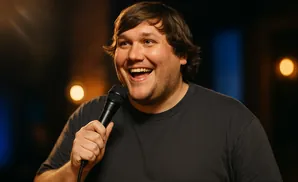 A man with brown hair and a gray t-shirt holds a microphone and laughs.