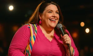 A woman with brown hair laughs while holding a microphone, wearing a pink shirt with a rainbow strap and pink earrings.