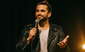 A man in a black leather jacket holds a microphone and gestures while speaking.