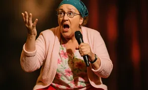A woman with glasses and a bandana on her head speaks into a microphone, gesturing with her left hand.