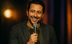 A man in a dark jacket and green shirt smiles into a microphone on stage.