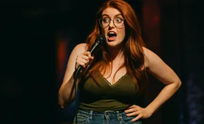 A surprised redhead woman with glasses holds a microphone onstage.