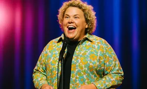 A woman with curly blonde hair, wearing a floral shirt, laughs into a microphone on stage with colorful lighting.