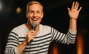 A man with blonde hair smiles and holds a microphone while raising his hand.