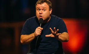 A man with a surprised expression holds a microphone and gestures with his hand.